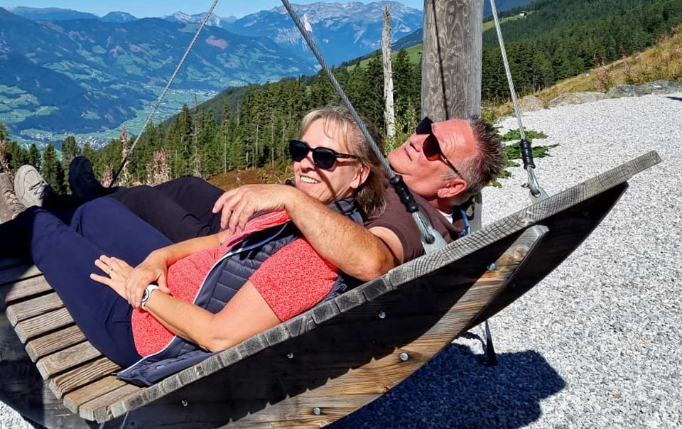 Die Gastgeber des Ferienhauses Casa La Vie stehen im Mittelpunkt des Bildes. Sie wirken herzlich und offen – ein echtes Herz für Gäste. Ihre entspannte Haltung und ihre freundlichen Gesichter zeigen gelebte Gastfreundschaft. Sie liegen gemeinsam auf einer Schaukel. Ihr Blick geht über ein Tal und auf Berge in der Ferne. Sie lachen und zeigen Humor und Entspannung. Ihre Präsenz vermittelt Vertrauen und lässt Gäste sich willkommen fühlen.