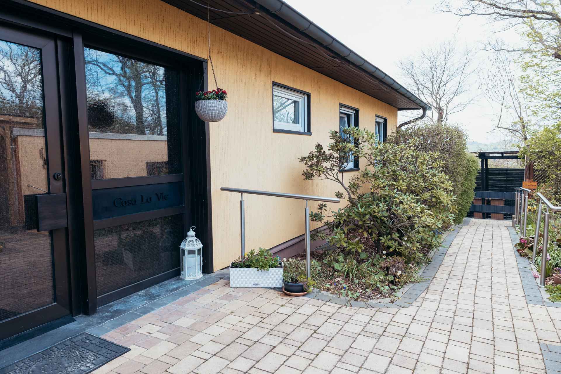 Barrierearmer Zugangsweg zur Ferienwohnung „Büdingen“ im Casa La Vie. Der gepflasterte Weg mit einseitigem Handlauf führt entlang des Hauses zum Eingangsbereich und Garten. Vor der Haustür befindet sich eine kleine Gummirampe, damit die Türschwelle leichter überfahren werden kann. Pflanzen, Blumen und helle Fassadenflächen sorgen für eine freundliche und gepflegte Atmosphäre.