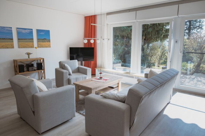 Helles, barrierearmes Wohnzimmer der Ferienwohnung „Büdingen“ im Casa La Vie in Altenstadt-Lindheim. Große Fensterflächen mit Blick auf Terrasse und Garten lassen viel Tageslicht in den Raum. Eine helle Sitzecke mit grauem Sofa, zwei Sesseln und Holzcouchtisch schafft eine ruhige und gemütliche Atmosphäre. Zur Ausstattung gehören außerdem ein Fernseher, Landschaftsbilder an der Wand sowie ein kleines Regal mit Musikanlage. Das Wohnzimmer wirkt modern, offen und freundlich gestaltet.