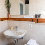 Das helle Badezimmer der Ferienwohnung „Keltenwelt“ in der Wetterau bietet Tageslicht durch ein Dachfenster, beigen Wandfliesen und moderne Ausstattung. Ein großer Spiegel mit moderner Beleuchtung hängt über einer Holzkonsole. Darunter befindet sich ein beigefarbenes Waschbecken. Links steht ein Edelstahl-Mülleimer, rechts ein schwarzer Hocker mit gefalteten weißen Handtüchern. Eine kleine Zimmerpflanze ergänzt die gepflegte und funktionale Einrichtung.