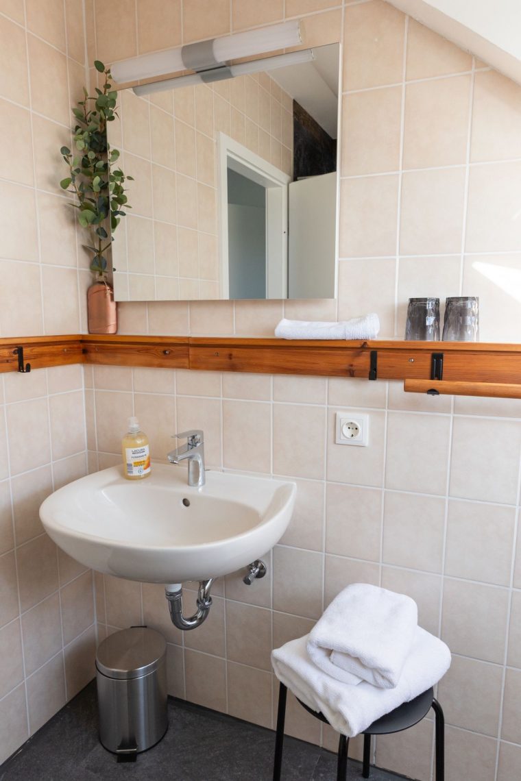 Das helle Badezimmer der Ferienwohnung „Keltenwelt“ in der Wetterau bietet Tageslicht durch ein Dachfenster, beigen Wandfliesen und moderne Ausstattung. Ein großer Spiegel mit moderner Beleuchtung hängt über einer Holzkonsole. Darunter befindet sich ein beigefarbenes Waschbecken. Links steht ein Edelstahl-Mülleimer, rechts ein schwarzer Hocker mit gefalteten weißen Handtüchern. Eine kleine Zimmerpflanze ergänzt die gepflegte und funktionale Einrichtung.