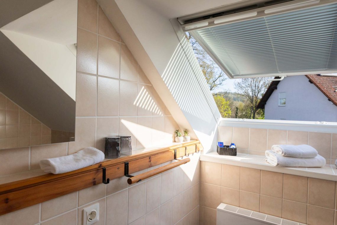 Die Ferienwohnung Keltenwelt bietet ein modernes Bad mit Tageslicht, Gartenblick, beigen Fliesen, Holzdetails, Handtüchern, Pflegeprodukten und Pflanzen. Ein großes Fenster mit Jalousie lässt viel Licht in den Raum. Die Wände sind mit beigen Fliesen verkleidet. Ein Handtuchhalter aus Holz ist an der Wand befestigt. Auf der Fensterbank liegen gefaltete Handtücher neben einem Korb mit Pflegeprodukten. Zimmerpflanzen sind im Raum verteilt und sorgen für eine angenehme Atmosphäre. Die Einrichtung ist zeitgemäß, durchdacht und einladend.