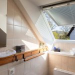 Die Ferienwohnung Keltenwelt bietet ein modernes Bad mit Tageslicht, Gartenblick, beigen Fliesen, Holzdetails, Handtüchern, Pflegeprodukten und Pflanzen. Ein großes Fenster mit Jalousie lässt viel Licht in den Raum. Die Wände sind mit beigen Fliesen verkleidet. Ein Handtuchhalter aus Holz ist an der Wand befestigt. Auf der Fensterbank liegen gefaltete Handtücher neben einem Korb mit Pflegeprodukten. Zimmerpflanzen sind im Raum verteilt und sorgen für eine angenehme Atmosphäre. Die Einrichtung ist zeitgemäß, durchdacht und einladend.