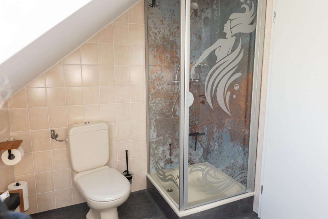 Die Ferienwohnung Keltenwelt bietet ein helles Bad mit Tageslicht, beigen Fliesen, gläserner Duschkabine, Wassernymphen-Motiv, Holzdetails und Gartenblick. Ein großes Dachfenster mit Jalousie lässt Tageslicht in den Raum und ermöglicht einen Blick ins Grüne. Die Wände sind mit beigen Fliesen verkleidet. Eine Duschkabine mit gläserner Tür zeigt das Motiv einer Wassernymphe mit fließendem Haar. Links daneben steht eine beigefarbene Standtoilette. Ein Toilettenpapierhalter aus Holz ist an der Wand befestigt. Die Einrichtung ist durchdacht, zeitgemäß und einladend.