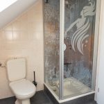 Die Ferienwohnung Keltenwelt bietet ein helles Bad mit Tageslicht, beigen Fliesen, gläserner Duschkabine, Wassernymphen-Motiv, Holzdetails und Gartenblick. Ein großes Dachfenster mit Jalousie lässt Tageslicht in den Raum und ermöglicht einen Blick ins Grüne. Die Wände sind mit beigen Fliesen verkleidet. Eine Duschkabine mit gläserner Tür zeigt das Motiv einer Wassernymphe mit fließendem Haar. Links daneben steht eine beigefarbene Standtoilette. Ein Toilettenpapierhalter aus Holz ist an der Wand befestigt. Die Einrichtung ist durchdacht, zeitgemäß und einladend.