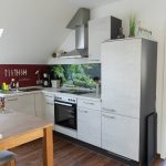 Die Küche der Ferienwohnung „Keltenwelt“ in der Wetterau bietet helle Arbeitsflächen, Herd, Backofen, Spüle, Dunstabzug und viel Tageslicht. Ein großes Fenster bringt Licht in den Raum. Rechts neben der Spüle steht eine Kaffeemaschine mit Dauerfilter. Ein kleiner Esstisch mit Blumenstrauß lädt zum Sitzen ein. An der roten Wand über der Spüle sind weiße keltische Buchstaben gut sichtbar. Ein Wandbild mit grünen Pflanzen schützt den Bereich über dem Herd vor Spritzern. Der angrenzende Wohnbereich mit Sofa und bodentiefen Fenstern ist direkt verbunden.