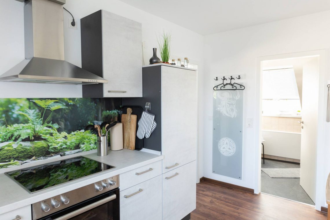 Die Küche der Ferienwohnung „Keltenwelt“ in der Wetterau bietet Cerankochfeld, Dunstabzug, helle Arbeitsfläche und Blick auf ein Naturmotiv. Grüne Pflanzen und Moos sind auf der Küchenrückwand abgebildet. Kochutensilien sind griffbereit. Rechts daneben liegt ein Badezimmer mit offener Tür. Eine Garderobe und ein Glasbild mit keltischem Muster ergänzen die Einrichtung. Die Küche lädt Gäste dazu ein, ihre Auszeit mit Komfort und Ruhe zu genießen