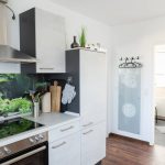 Die Küche der Ferienwohnung „Keltenwelt“ in der Wetterau bietet Cerankochfeld, Dunstabzug, helle Arbeitsfläche und Blick auf ein Naturmotiv. Grüne Pflanzen und Moos sind auf der Küchenrückwand abgebildet. Kochutensilien sind griffbereit. Rechts daneben liegt ein Badezimmer mit offener Tür. Eine Garderobe und ein Glasbild mit keltischem Muster ergänzen die Einrichtung. Die Küche lädt Gäste dazu ein, ihre Auszeit mit Komfort und Ruhe zu genießen