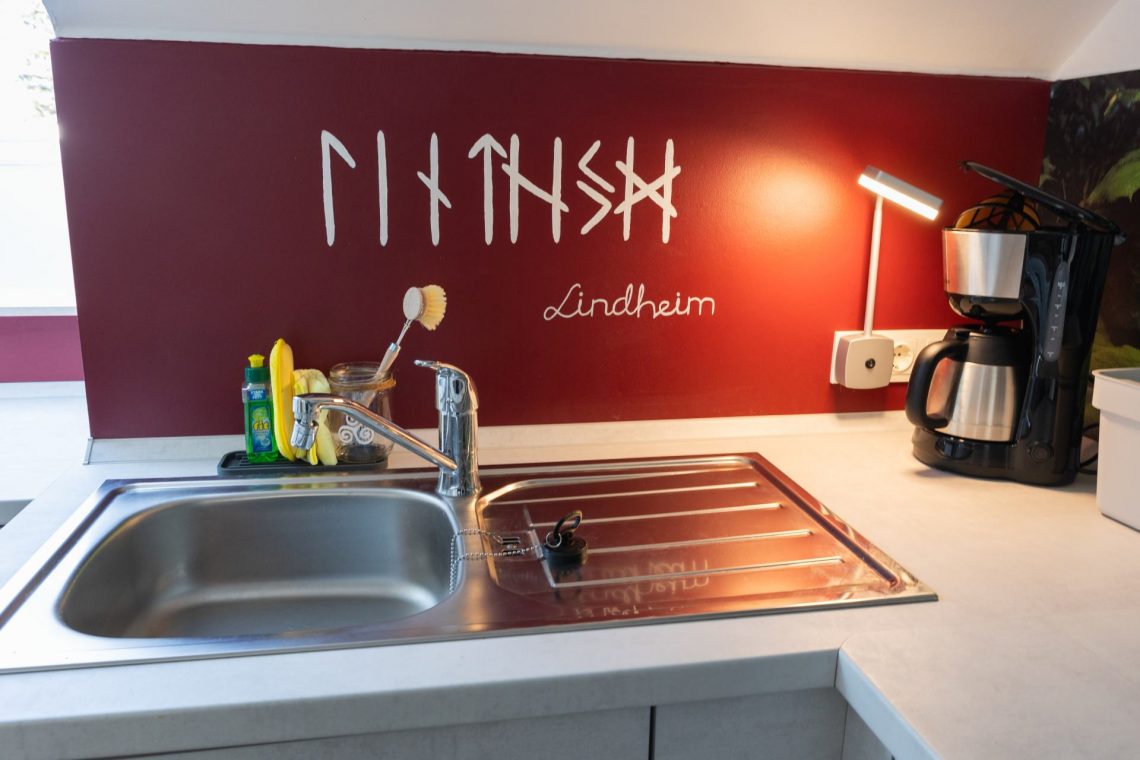 Die Küche der Ferienwohnung „Keltenwelt“ bietet Spüle, Kaffeemaschine mit Thermoskanne, rotes Wandmotiv mit Ornamenten und viel Tageslicht. Die Küche der Ferienwohnung „Keltenwelt“ bietet eine chromfarbene Spüle und eine Kaffeemaschine mit Permanentfilter und Thermoskanne. Die Wand hinter der Spüle ist rot gestrichen und zeigt keltische Ornamente sowie den Schriftzug „Lindheim“. Tageslicht fällt durch ein großes Dachfenster. Eine Deckenlampe und eine Steckdosenlampe sorgen für zusätzliche Helligkeit. Die offene Raumgestaltung erleichtert die Orientierung. Die Einrichtung verbindet moderne Funktionalität mit regionalem Design.