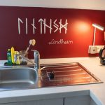 Die Küche der Ferienwohnung „Keltenwelt“ bietet Spüle, Kaffeemaschine mit Thermoskanne, rotes Wandmotiv mit Ornamenten und viel Tageslicht. Die Küche der Ferienwohnung „Keltenwelt“ bietet eine chromfarbene Spüle und eine Kaffeemaschine mit Permanentfilter und Thermoskanne. Die Wand hinter der Spüle ist rot gestrichen und zeigt keltische Ornamente sowie den Schriftzug „Lindheim“. Tageslicht fällt durch ein großes Dachfenster. Eine Deckenlampe und eine Steckdosenlampe sorgen für zusätzliche Helligkeit. Die offene Raumgestaltung erleichtert die Orientierung. Die Einrichtung verbindet moderne Funktionalität mit regionalem Design.