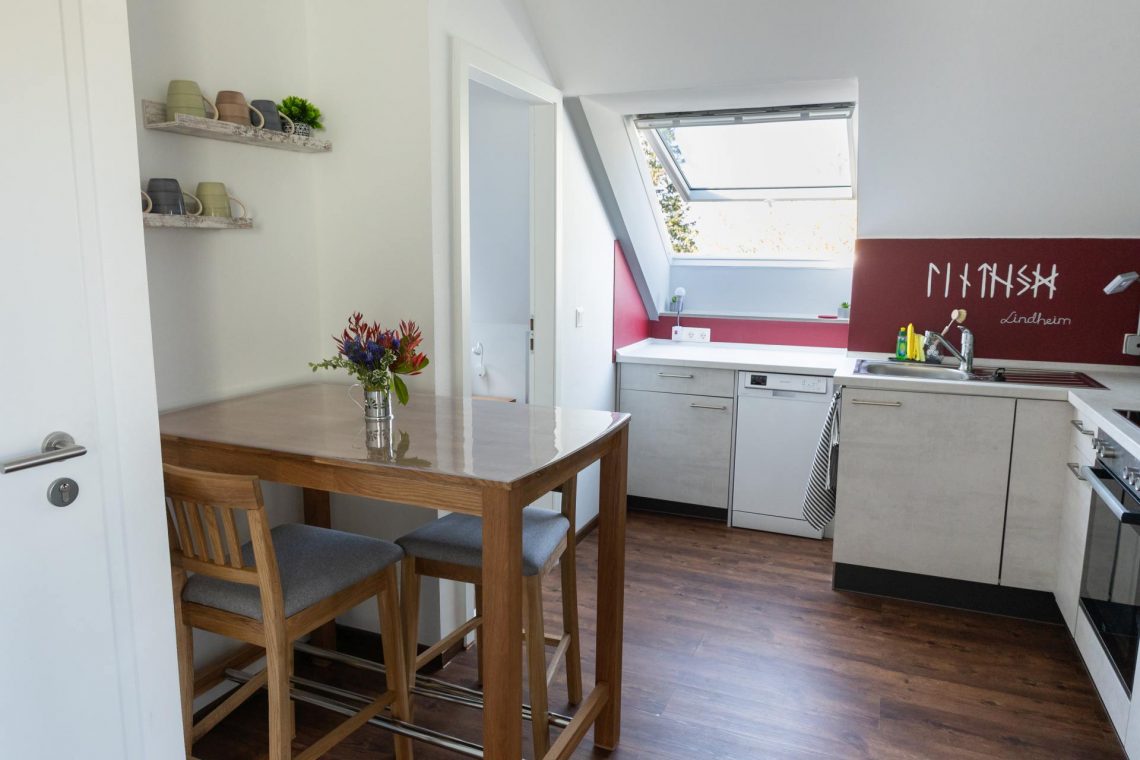 Die Küche der Ferienwohnung „Keltenwelt“ zeigt eine helle Küchenzeile mit weißen Fronten. Sie enthält eine Spüle, einen Geschirrspüler und einen Backofen. Über der Spüle ist der Schriftzug „Lindheim“ mit keltischem Bezug angebracht. Vor der Küchenzeile steht ein Holz-Esstisch mit einem Blumenarrangement. Ein Wandregal mit Kaffeetassen befindet sich in der Nähe. Der Boden ist dunkel und holzfarben. Eine kleine Pflanze ergänzt die Einrichtung. Die Küche ist funktional und kombiniert modernes Design mit regionalen Elementen.