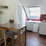 Die Küche der Ferienwohnung „Keltenwelt“ zeigt eine helle Küchenzeile mit weißen Fronten. Sie enthält eine Spüle, einen Geschirrspüler und einen Backofen. Über der Spüle ist der Schriftzug „Lindheim“ mit keltischem Bezug angebracht. Vor der Küchenzeile steht ein Holz-Esstisch mit einem Blumenarrangement. Ein Wandregal mit Kaffeetassen befindet sich in der Nähe. Der Boden ist dunkel und holzfarben. Eine kleine Pflanze ergänzt die Einrichtung. Die Küche ist funktional und kombiniert modernes Design mit regionalen Elementen.