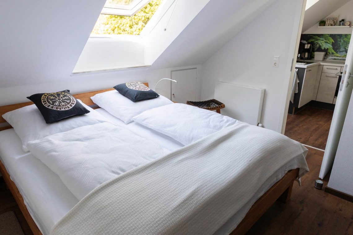Die Ferienwohnung Keltenwelt verfügt über ein helles Schlafzimmer mit Eichenbett, weißer Bettwäsche, keltischen Dekokissen und moderner Ausstattung. Ein großes Dachfenster lässt Tageslicht in den Raum. Im Hintergrund befindet sich eine offene Küche. Die Einrichtung ist modern und gut nutzbar.