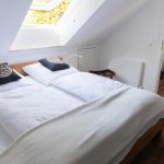 Die Ferienwohnung Keltenwelt verfügt über ein helles Schlafzimmer mit Eichenbett, weißer Bettwäsche, keltischen Dekokissen und moderner Ausstattung. Ein großes Dachfenster lässt Tageslicht in den Raum. Im Hintergrund befindet sich eine offene Küche. Die Einrichtung ist modern und gut nutzbar.
