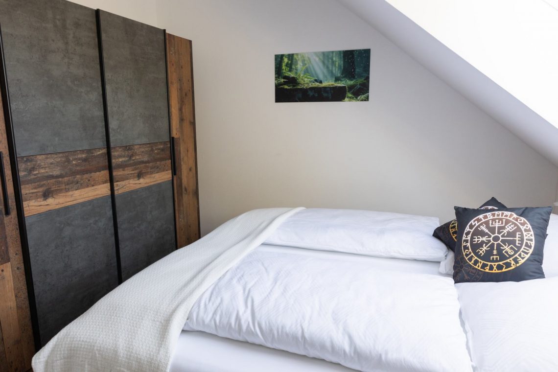 Ferienwohnung Keltenwelt – Helles Schlafzimmer mit Eichenbett und keltischen Details. Das Schlafzimmer der Ferienwohnung „Keltenwelt“ in der Wetterau liegt unter einer Dachschräge. Ein großes Dachfenster lässt Tageslicht in den Raum. Ein Doppelbett mit weißer Bettwäsche steht mittig im Zimmer. Schwarze Dekokissen mit keltischen Symbolen liegen auf dem Bett. Ein rustikaler Kleiderschrank in Holzoptik befindet sich gegenüber. An der Wand hängt ein Bild mit Sonnenstrahlen, die durch einen Wald fallen. Die Einrichtung ist harmonisch abgestimmt, wohnlich und gut durchdacht.