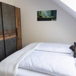 Ferienwohnung Keltenwelt – Helles Schlafzimmer mit Eichenbett und keltischen Details. Das Schlafzimmer der Ferienwohnung „Keltenwelt“ in der Wetterau liegt unter einer Dachschräge. Ein großes Dachfenster lässt Tageslicht in den Raum. Ein Doppelbett mit weißer Bettwäsche steht mittig im Zimmer. Schwarze Dekokissen mit keltischen Symbolen liegen auf dem Bett. Ein rustikaler Kleiderschrank in Holzoptik befindet sich gegenüber. An der Wand hängt ein Bild mit Sonnenstrahlen, die durch einen Wald fallen. Die Einrichtung ist harmonisch abgestimmt, wohnlich und gut durchdacht.