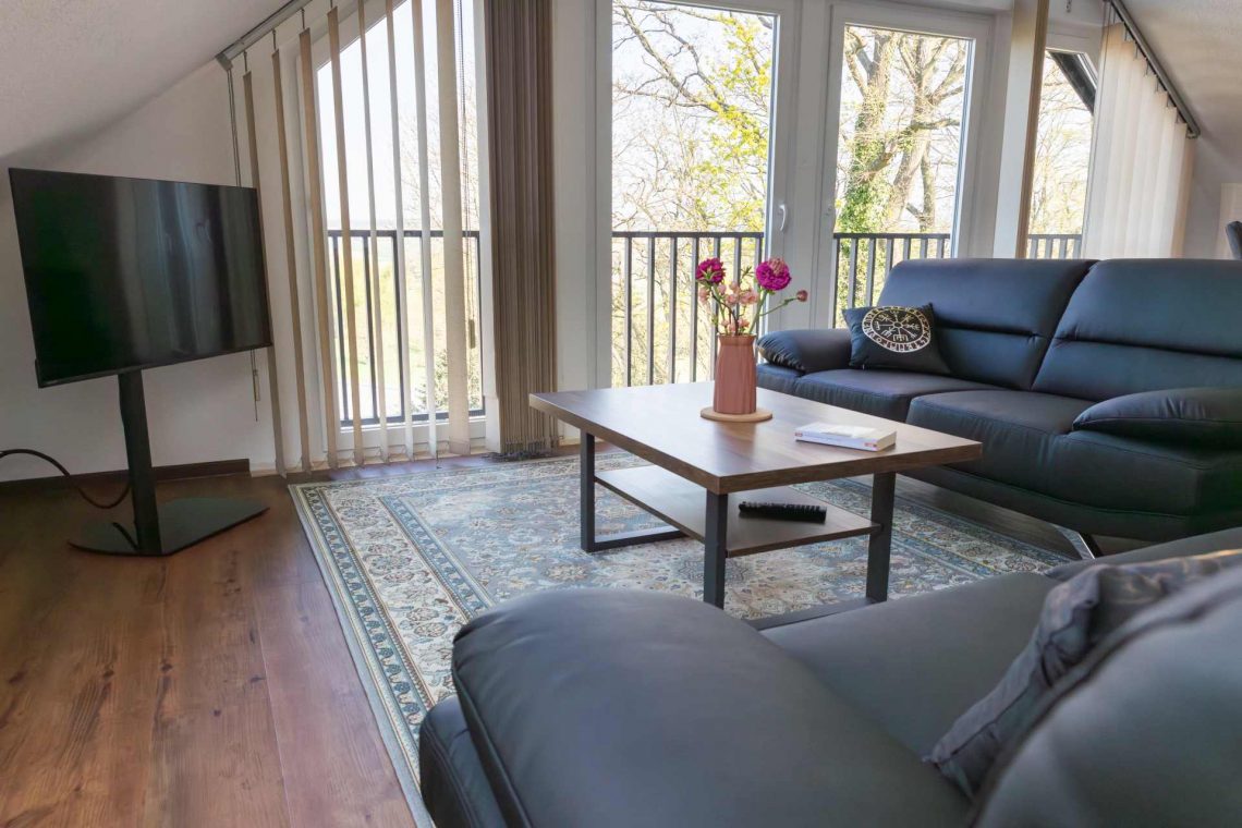 Helles Wohnzimmer im Dachgeschoss der Ferienwohnung „Keltenwelt“ in der Wetterau. Große Fenster lassen viel Tageslicht in den Raum und bieten einen Panoramablick ins Grüne sowie auf drei Storchennester. Zwei schwarze Ledersofas stehen gegenüber einem rustikalen Holztisch, auf dem ein bunter Blumenstrauß arrangiert ist. In der linken Zimmerecke befindet sich ein moderner Fernseher. Dekorationselemente sind im Raum verteilt. Die Einrichtung ist geschmackvoll und alltagstauglich gestaltet.