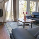 Helles Wohnzimmer im Dachgeschoss der Ferienwohnung „Keltenwelt“ in der Wetterau. Große Fenster lassen viel Tageslicht in den Raum und bieten einen Panoramablick ins Grüne sowie auf drei Storchennester. Zwei schwarze Ledersofas stehen gegenüber einem rustikalen Holztisch, auf dem ein bunter Blumenstrauß arrangiert ist. In der linken Zimmerecke befindet sich ein moderner Fernseher. Dekorationselemente sind im Raum verteilt. Die Einrichtung ist geschmackvoll und alltagstauglich gestaltet.