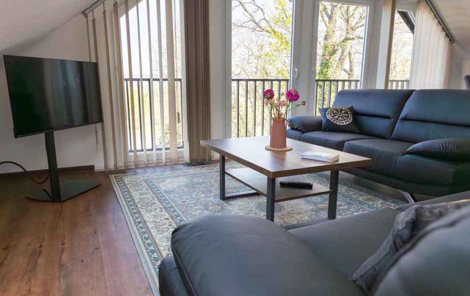 Helles Wohnzimmer im Dachgeschoss der Ferienwohnung „Keltenwelt“ in der Wetterau. Große Fenster lassen viel Tageslicht in den Raum und bieten einen Panoramablick ins Grüne sowie auf drei Storchennester. Zwei schwarze Ledersofas stehen gegenüber einem rustikalen Holztisch, auf dem ein bunter Blumenstrauß arrangiert ist. In der linken Zimmerecke befindet sich ein moderner Fernseher. Dekorationselemente sind im Raum verteilt. Die Einrichtung ist geschmackvoll und alltagstauglich gestaltet.