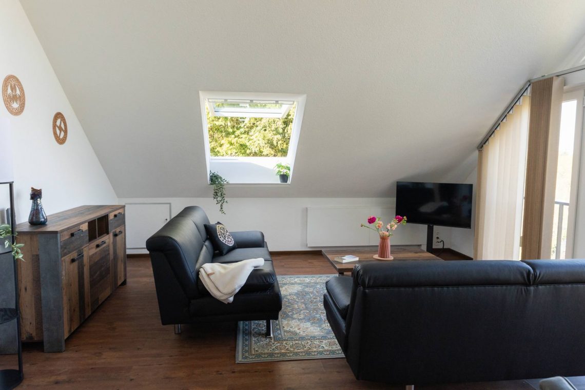 Helles großes Wohnzimmer im Dachgeschoss der Ferienwohnung Keltenwelt. Zwei schwarze Ledersofas stehen gegenüber einem rustikalen Holztisch, auf dem ein bunter Blumenstrauß arrangiert ist. Auf dem linken Sofa liegt eine helle Wolldecke. Ein großzügiger Fensterbereich lässt viel Tageslicht in den Raum und bietet Blick ins Grüne. In der rechten hinteren Zimmerecke steht ein moderner Fernseher. Die Einrichtung ist gemütlich und funktional – geeignet für Urlaub, Arbeitspausen oder kurze Auszeiten.