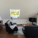 Helles großes Wohnzimmer im Dachgeschoss der Ferienwohnung Keltenwelt. Zwei schwarze Ledersofas stehen gegenüber einem rustikalen Holztisch, auf dem ein bunter Blumenstrauß arrangiert ist. Auf dem linken Sofa liegt eine helle Wolldecke. Ein großzügiger Fensterbereich lässt viel Tageslicht in den Raum und bietet Blick ins Grüne. In der rechten hinteren Zimmerecke steht ein moderner Fernseher. Die Einrichtung ist gemütlich und funktional – geeignet für Urlaub, Arbeitspausen oder kurze Auszeiten.