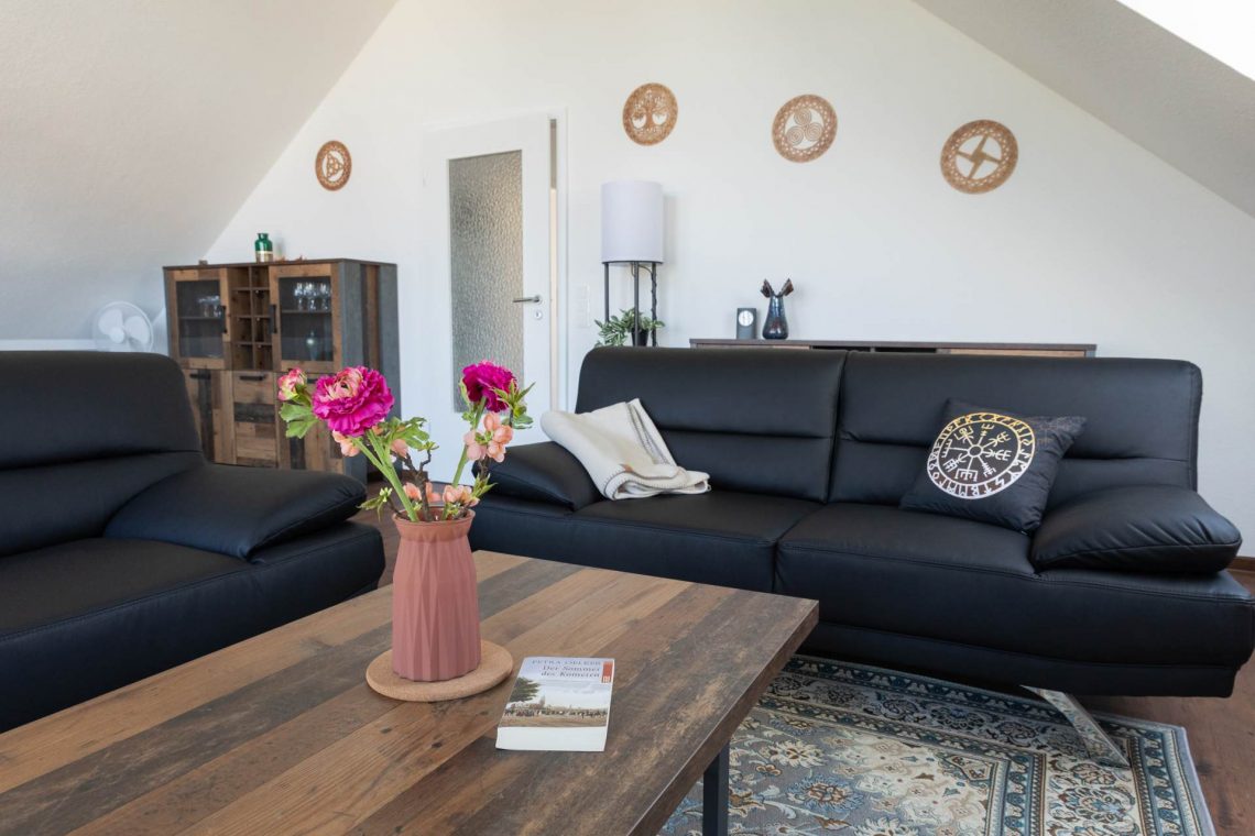 Großes, helles Wohnzimmer im Dachgeschoss der Ferienwohnung Keltenwelt. Zwei moderne Ledersofas stehen gegenüber einem rustikalen Holztisch, auf dem ein bunter Blumenstrauß arrangiert ist. Keltische Wandmotive und ein elegantes Sideboard ergänzen die Einrichtung. Großzügige Fensterflächen lassen viel Tageslicht in den Raum und bieten Blick ins Grüne. Die Einrichtung wirkt aufgeräumt und individuell gestaltet.