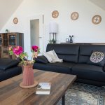 Großes, helles Wohnzimmer im Dachgeschoss der Ferienwohnung Keltenwelt. Zwei moderne Ledersofas stehen gegenüber einem rustikalen Holztisch, auf dem ein bunter Blumenstrauß arrangiert ist. Keltische Wandmotive und ein elegantes Sideboard ergänzen die Einrichtung. Großzügige Fensterflächen lassen viel Tageslicht in den Raum und bieten Blick ins Grüne. Die Einrichtung wirkt aufgeräumt und individuell gestaltet.