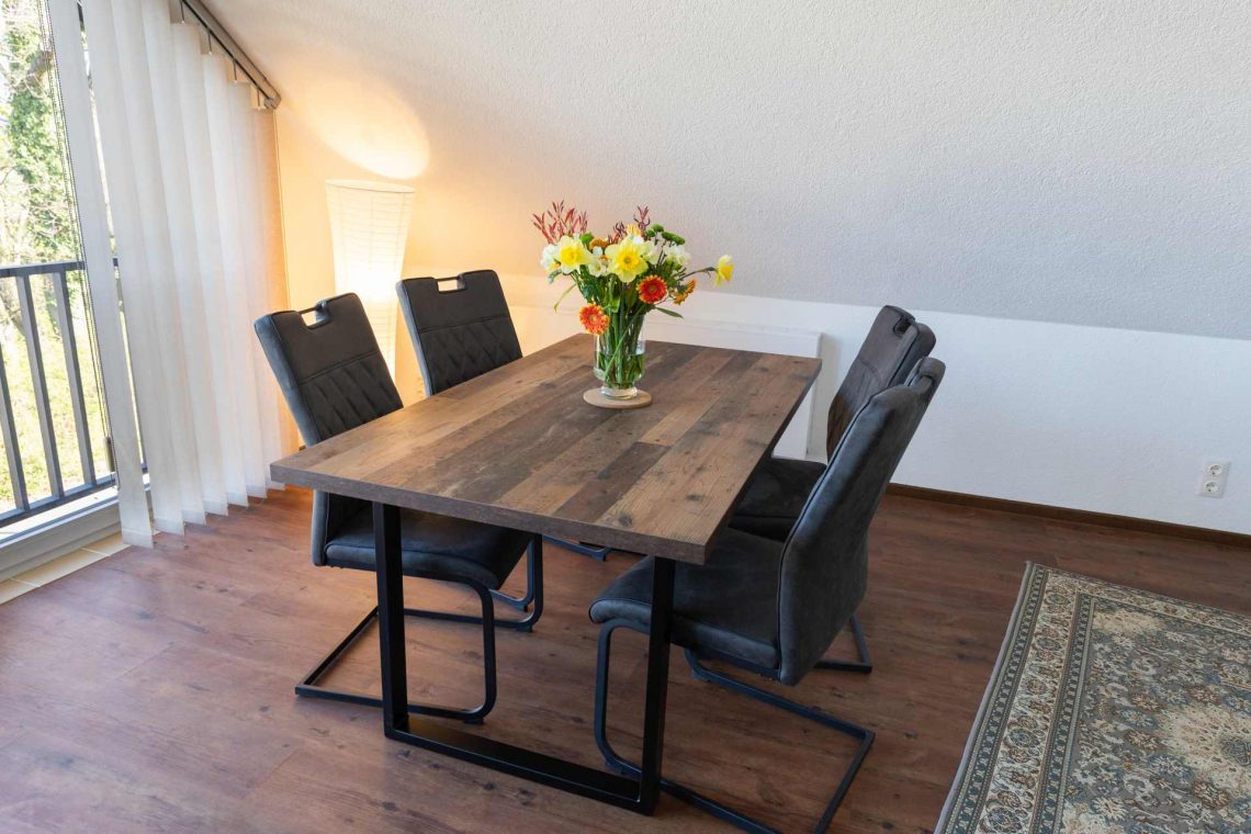 Lichtdurchfluteter Essbereich im Dachgeschoss der Ferienwohnung Keltenwelt. Ein großzügiger Esstisch mit vier Stühlen steht in einem aufgeräumten Raum. Große Fenster mit Vertikaljalousien lassen viel Tageslicht hinein und bieten Blick ins Grüne. Das offene Raumkonzept wirkt modern und funktional.