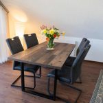 Lichtdurchfluteter Essbereich im Dachgeschoss der Ferienwohnung Keltenwelt. Ein großzügiger Esstisch mit vier Stühlen steht in einem aufgeräumten Raum. Große Fenster mit Vertikaljalousien lassen viel Tageslicht hinein und bieten Blick ins Grüne. Das offene Raumkonzept wirkt modern und funktional.