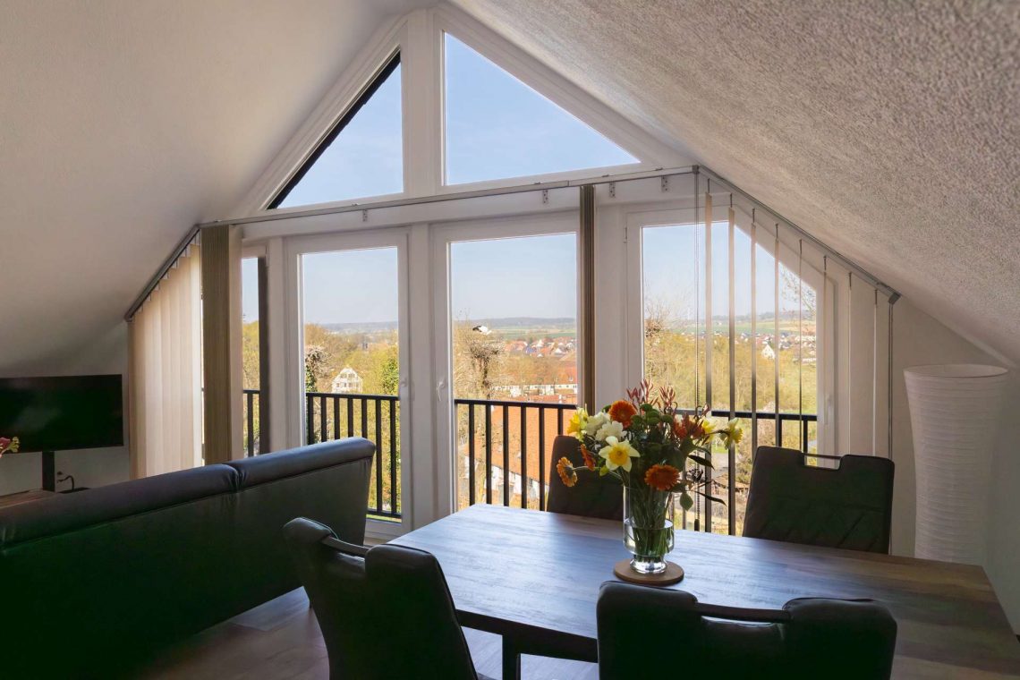 Offenes, großes Wohnzimmer im Dachgeschoss der Ferienwohnung „Keltenwelt“ in der Wetterau. Ein großzügiger Esstisch mit Blumenstrauß steht im Raum. Eine weiße Stehlampe befindet sich in der rechten Zimmerecke. Breite Fensterflächen lassen viel Tageslicht hinein und bieten Panoramablick auf drei Storchennester, das Dorf Lindheim und die umliegende Landschaft der Wetterau. Die Einrichtung ist stilvoll und aufgeräumt.