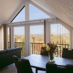 Offenes, großes Wohnzimmer im Dachgeschoss der Ferienwohnung „Keltenwelt“ in der Wetterau. Ein großzügiger Esstisch mit Blumenstrauß steht im Raum. Eine weiße Stehlampe befindet sich in der rechten Zimmerecke. Breite Fensterflächen lassen viel Tageslicht hinein und bieten Panoramablick auf drei Storchennester, das Dorf Lindheim und die umliegende Landschaft der Wetterau. Die Einrichtung ist stilvoll und aufgeräumt.