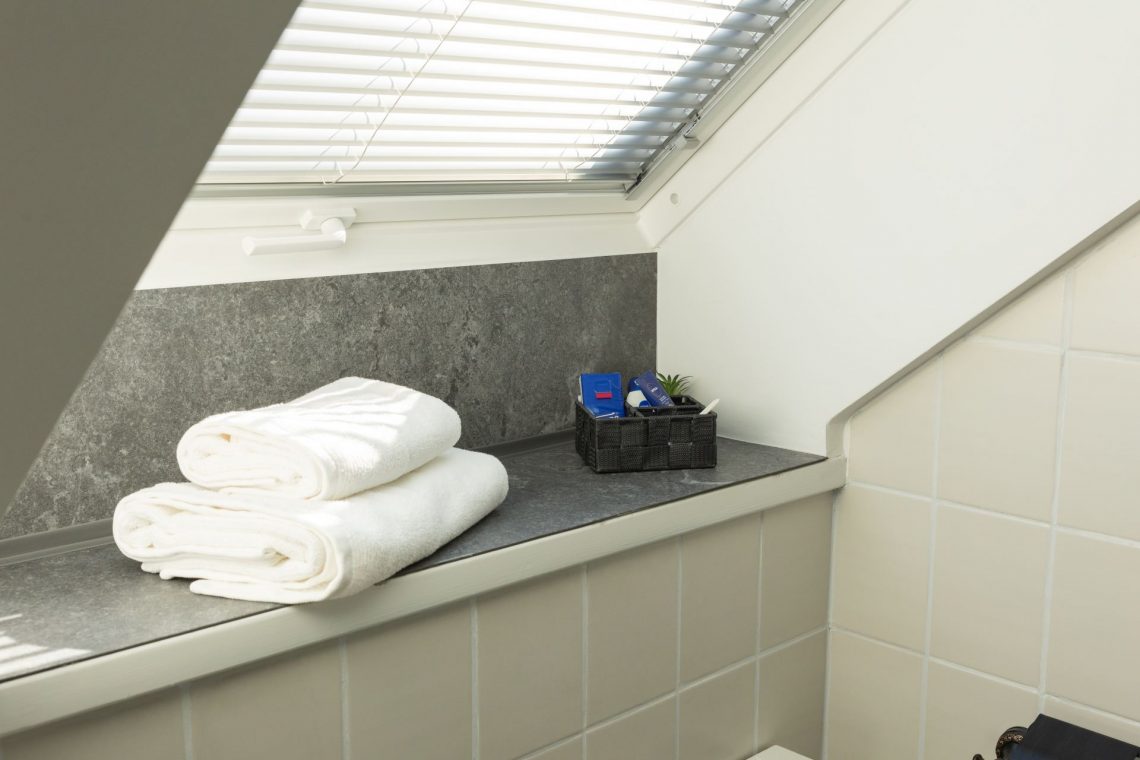 Helles Tageslichtbad mit Dachfenster in der Ferienwohnung „Ronneburg“. Durch das geöffnete Fenster fällt der Blick ins Grüne. Jalousien ermöglichen eine flexible Lichtregulierung. Auf der breiten Fensterbank liegen zwei gefaltete weiße Handtücher und ein schwarzer Korb mit Pflegeprodukten. Die Einrichtung wirkt aufgeräumt und stilvoll. Das natürliche Tageslicht sorgt für eine freundliche Atmosphäre.