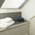 Helles Tageslichtbad mit Dachfenster in der Ferienwohnung „Ronneburg“. Durch das geöffnete Fenster fällt der Blick ins Grüne. Jalousien ermöglichen eine flexible Lichtregulierung. Auf der breiten Fensterbank liegen zwei gefaltete weiße Handtücher und ein schwarzer Korb mit Pflegeprodukten. Die Einrichtung wirkt aufgeräumt und stilvoll. Das natürliche Tageslicht sorgt für eine freundliche Atmosphäre.