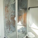 Stilvolles Duschbad in der Ferienwohnung „Ronneburg“. Die transparente Duschabtrennung zeigt eine Illustration einer Wassernymphe mit langem Haar. Die rückwärtige Wand hat eine rustikal strukturierte Oberfläche. Neben der Dusche steht ein weißer Hocker mit einem gefalteten weißen Handtuch. An der Wand hängt ein Handtuchhalter mit einem weiteren weißen Handtuch. Die Einrichtung wirkt ruhig und aufgeräumt.