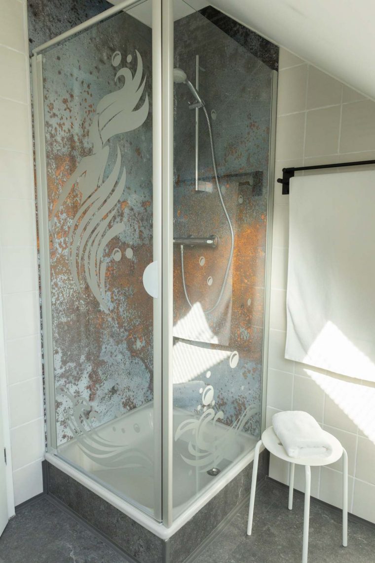 Stilvolles Duschbad in der Ferienwohnung „Ronneburg“. Die transparente Duschabtrennung zeigt eine Illustration einer Wassernymphe mit langem Haar. Die rückwärtige Wand hat eine rustikal strukturierte Oberfläche. Neben der Dusche steht ein weißer Hocker mit einem gefalteten weißen Handtuch. An der Wand hängt ein Handtuchhalter mit einem weiteren weißen Handtuch. Die Einrichtung wirkt ruhig und aufgeräumt.