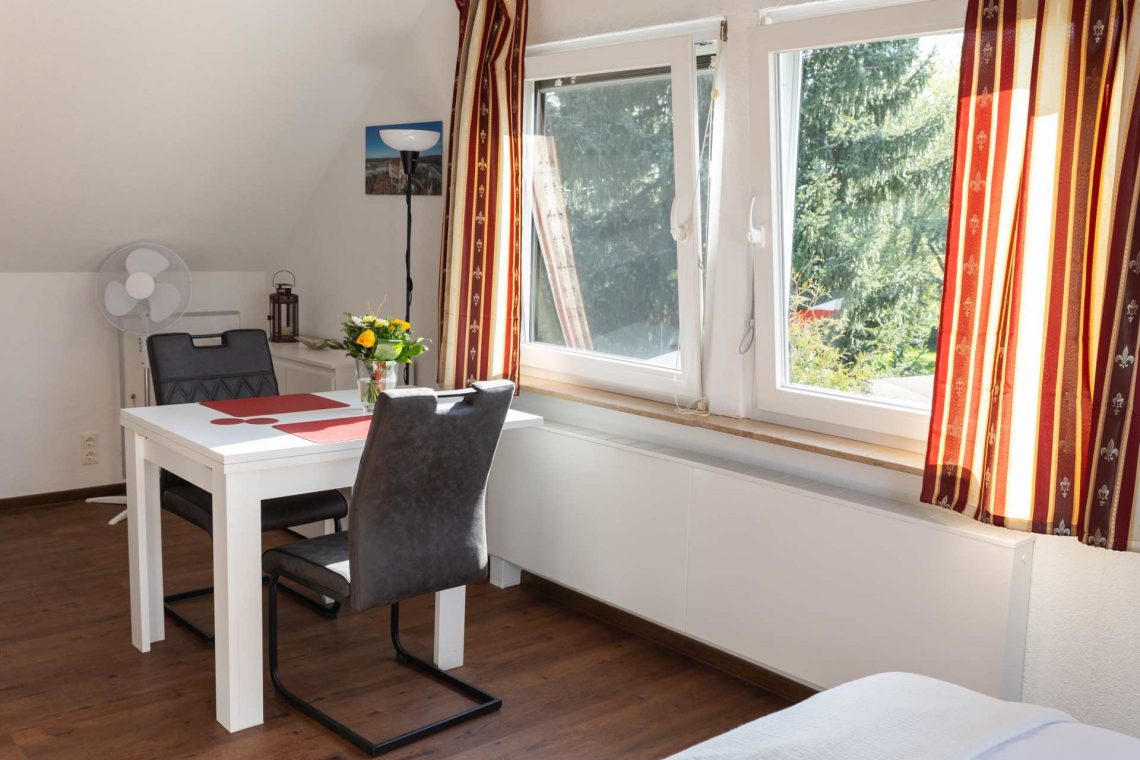 Heller Ess- und Wohnbereich in der Ferienwohnung „Ronneburg“. Ein großer, ausziehbarer Esstisch in Weiß mit roten Platzsets und einer Blumenvase steht im Raum. Zwei gepolsterte Stühle sind bereitgestellt. Große Fenster lassen viel Tageslicht hinein und bieten Blick auf die umliegende Natur. Ein elegantes Sideboard bietet Stauraum für Geschirr und persönliche Gegenstände. Auf dem Sideboard steht eine rote Laterne. In der linken Zimmerecke befindet ich ein Standventilator. Die Einrichtung wirkt aufgeräumt und stilvoll.