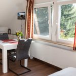 Heller Ess- und Wohnbereich in der Ferienwohnung „Ronneburg“. Ein großer, ausziehbarer Esstisch in Weiß mit roten Platzsets und einer Blumenvase steht im Raum. Zwei gepolsterte Stühle sind bereitgestellt. Große Fenster lassen viel Tageslicht hinein und bieten Blick auf die umliegende Natur. Ein elegantes Sideboard bietet Stauraum für Geschirr und persönliche Gegenstände. Auf dem Sideboard steht eine rote Laterne. In der linken Zimmerecke befindet ich ein Standventilator. Die Einrichtung wirkt aufgeräumt und stilvoll.