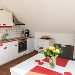 Helle Dachgeschossküche in der Ferienwohnung „Ronneburg“. Weiße Schränke mit roten Akzenten prägen den Raum. Zur Ausstattung gehören Edelstahlspüle, ein Kühlschrank, ein Ceranherd, eine Dunstabzugshaube und ein Backofen. Ein Esstisch mit roten Platzsets und einem Blumenstrauß steht mittig im Raum. Eine Kaffeemaschine mit Permanentfilter und Warmhaltekanne sowie diverse Kochutensilien befindet sich auf der Arbeitsfläche. Ein mobiler Servierwagen mit Fächern und Flaschenregalen steht daneben. Im Hintergrund sorgt ein Standventilator für Luftzirkulation. Die Einrichtung wirkt aufgeräumt und individuell gestaltet.