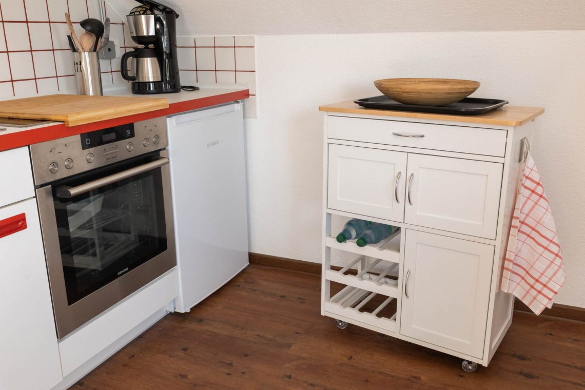 Liebevoll eingerichtete Dachgeschossküche in der Ferienwohnung „Ronneburg“. Die Ausstattung umfasst ein Ceranfeld, einen Backofen und einen Kühlschrank. Auf dem Kochfeld liegt ein Bambusbrett als zusätzliche Ablagefläche. Eine Kaffeemaschine mit Permanentfilter und Warmhaltekanne steht auf der Arbeitsfläche. Ein mobiler Servierwagen mit Schubladen und Flaschenregalen ergänzt die Einrichtung. Verschiedene Kochutensilien wie Pfannenwender, Holzlöffel und Kelle sind griffbereit. Die Küche wirkt funktional und stilvoll.