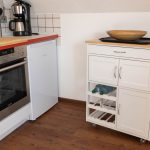 Liebevoll eingerichtete Dachgeschossküche in der Ferienwohnung „Ronneburg“. Die Ausstattung umfasst ein Ceranfeld, einen Backofen und einen Kühlschrank. Auf dem Kochfeld liegt ein Bambusbrett als zusätzliche Ablagefläche. Eine Kaffeemaschine mit Permanentfilter und Warmhaltekanne steht auf der Arbeitsfläche. Ein mobiler Servierwagen mit Schubladen und Flaschenregalen ergänzt die Einrichtung. Verschiedene Kochutensilien wie Pfannenwender, Holzlöffel und Kelle sind griffbereit. Die Küche wirkt funktional und stilvoll.