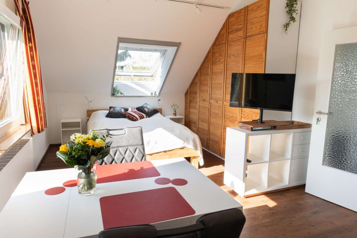 Helle Ferienwohnung mit großem Dachfenster und zwei weiteren Tageslichtfenstern mit Blick ins Grüne. Unter dem Dachfenster steht ein Doppelbett aus hellem Eichenholz. Ein mehrtüriger Holzeinbauschrank und mehrere Pflanzen verleihen dem Raum natürliche Akzente. Links neben der Tür befindet sich ein kleines weißes Regal mit einem modernen Fernseher, der über einen Schubtisch ausgefahren werden kann. Im Vordergrund steht ein ausziehbarer weißer Tisch mit roten Platzsets und einem Blumenstrauß. Zwei Polsterstühle stehen am Tisch. Die Einrichtung wirkt freundlich und charmant. Der Ausblick zeigt Bäume und Landschaft der Wetterau.