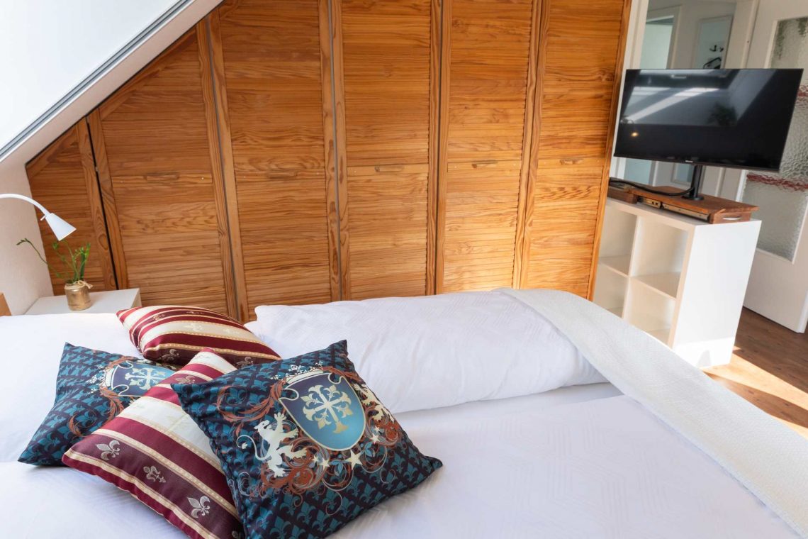 Schlafbereich in der Ferienwohnung Ronneburg mit breitem Doppelbett und stilvollen Kissen, darunter auch Kissen mit Wappenmotiv. Hinter dem Bett steht ein großer, holzfarbewner Einbauschrank mit mehreren Türen. Rechts daneben befindet sich ein kleines weißes Regal mit einem modernen Flachbildfernseher. Links sorgt ein Dachfenster für Tageslicht. Auf dem Nachttisch steht eine kleine Pflanze. Die Einrichtung wirkt gemütlich und charmant.