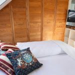 Schlafbereich in der Ferienwohnung Ronneburg mit breitem Doppelbett und stilvollen Kissen, darunter auch Kissen mit Wappenmotiv. Hinter dem Bett steht ein großer, holzfarbewner Einbauschrank mit mehreren Türen. Rechts daneben befindet sich ein kleines weißes Regal mit einem modernen Flachbildfernseher. Links sorgt ein Dachfenster für Tageslicht. Auf dem Nachttisch steht eine kleine Pflanze. Die Einrichtung wirkt gemütlich und charmant.